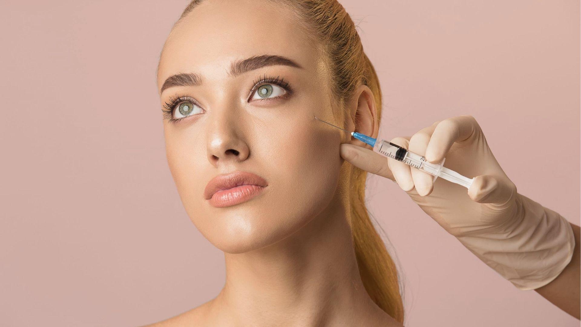 Young woman receiving cheek filler injection with syringe near cheekbone against pink background