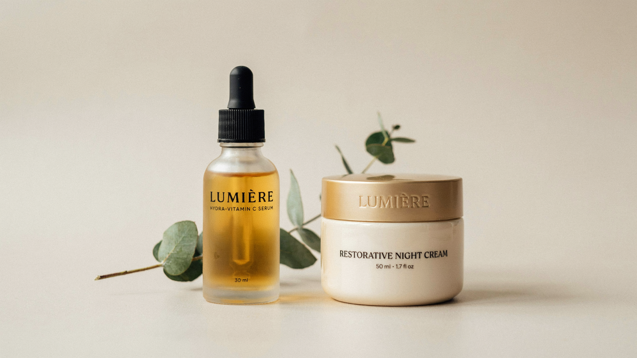 Lumière Hydra-Vitamin C Serum and Restorative Night Cream with gold lid arranged on a neutral background with eucalyptus.