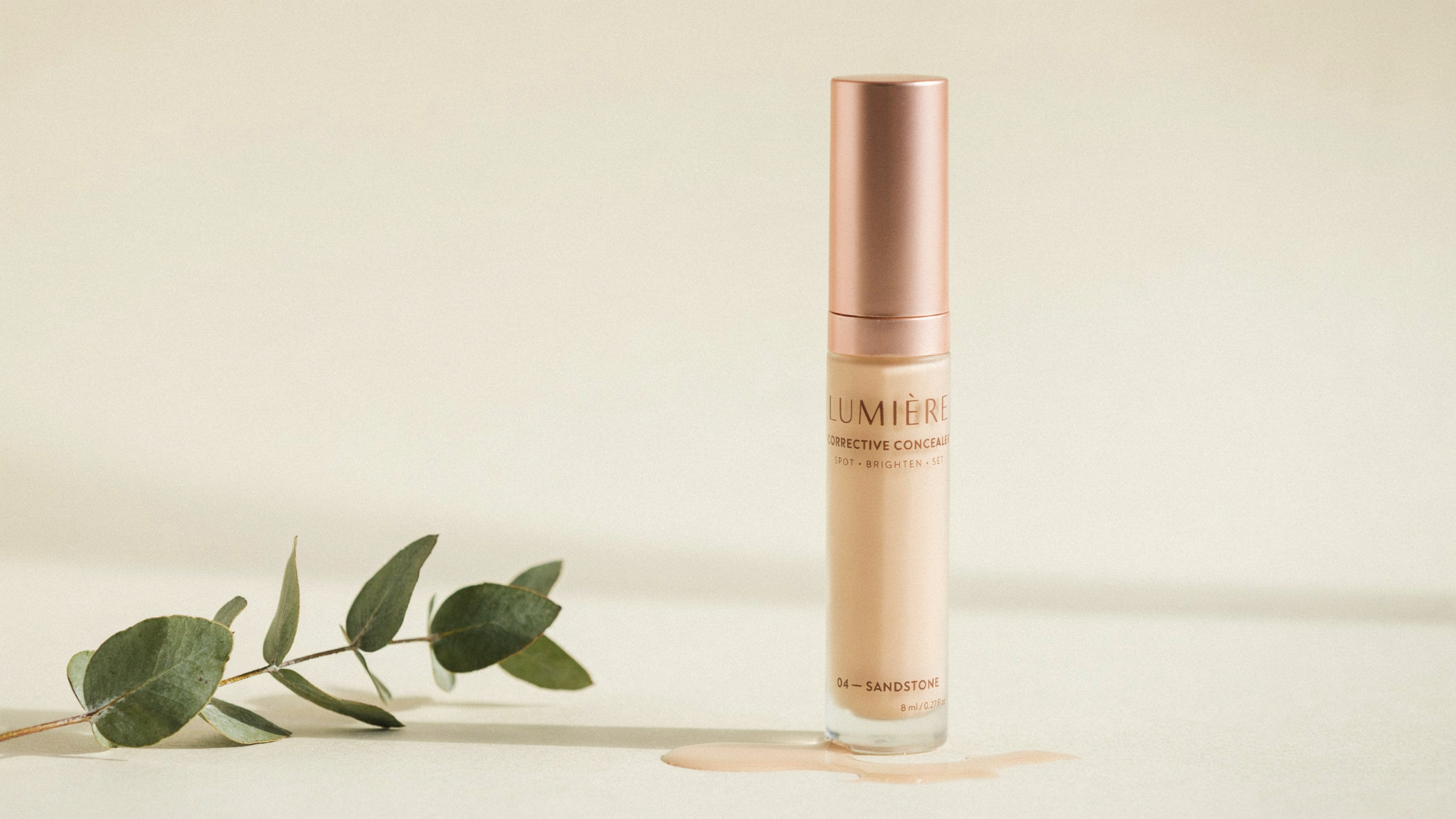 Lumière Corrective Concealer in shade 04 Sandstone with a rose gold cap, displayed beside a eucalyptus sprig on a beige surface.