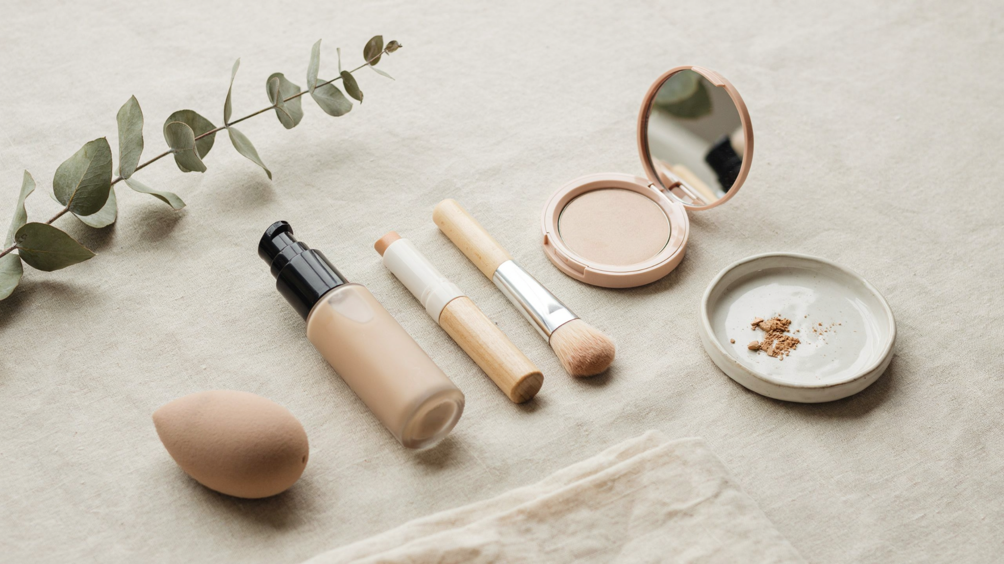 A beauty flat lay featuring a makeup sponge, liquid foundation, concealer stick.