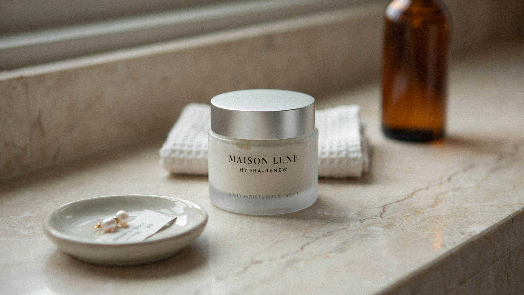 Maison Lune Hydra-Renew Daily Moisturizer frosted jar with silver lid on a marble countertop beside pearl earrings.