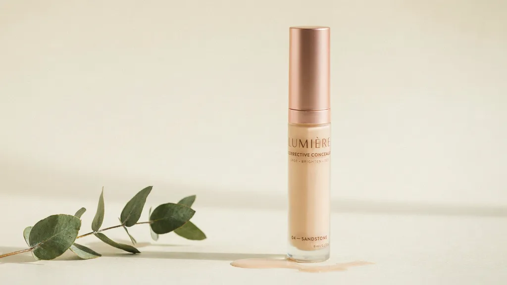 Lumière Corrective Concealer in shade 04 Sandstone with a rose gold cap, displayed beside a eucalyptus sprig on a beige surface.