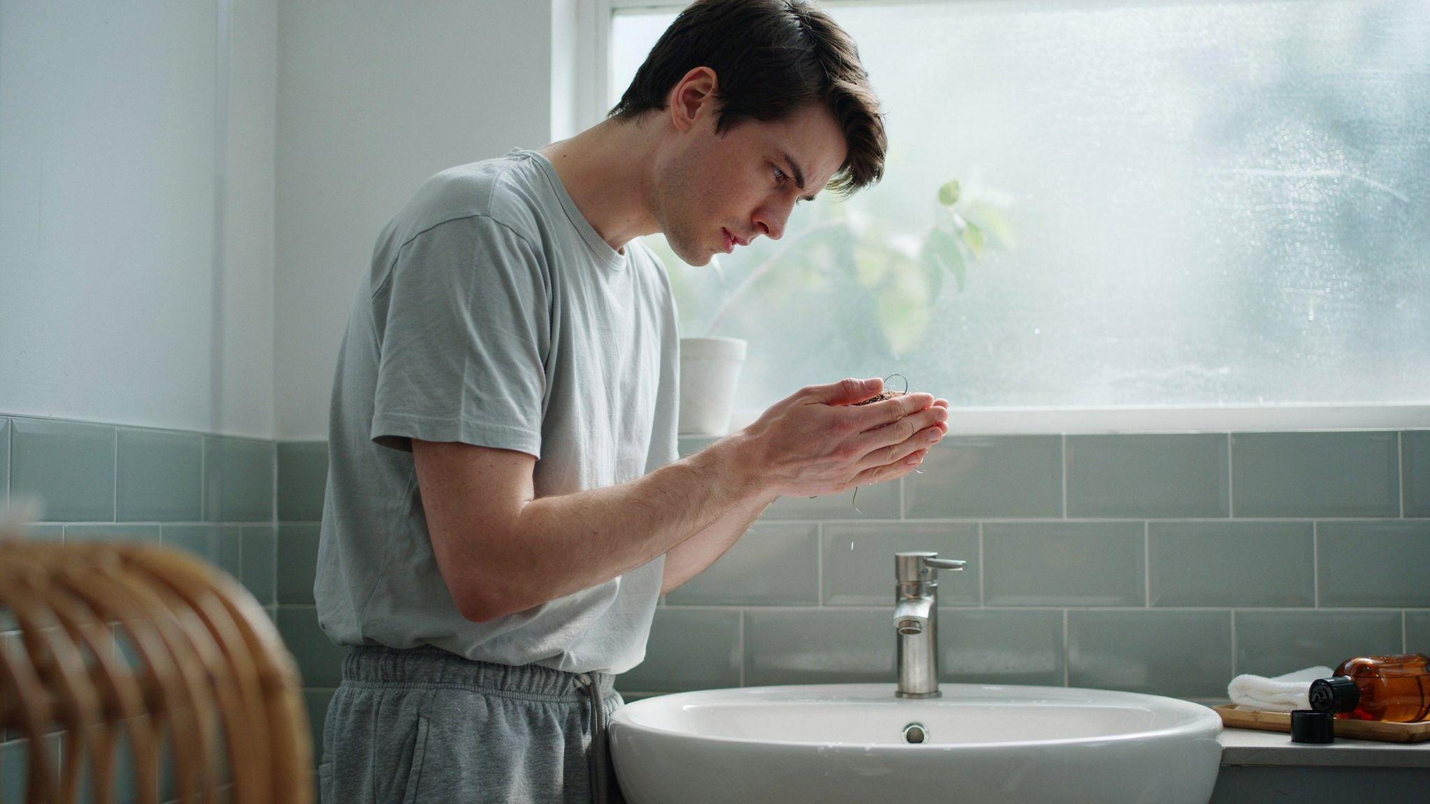 "Young adult in bathroom looking worried while holding shed hair in hand under natural light"