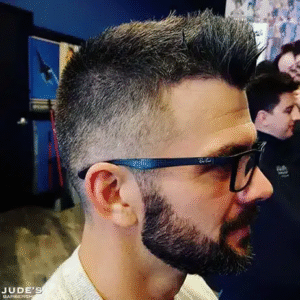 A man with glasses and a beard sits in a barber shop, preparing for a haircut.