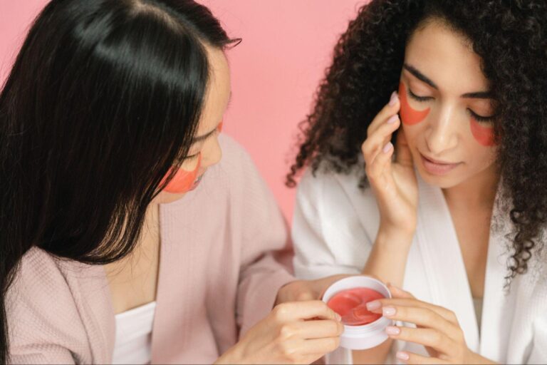 Two women are applying makeup to their faces, focusing on enhancing their features in a bright, well-lit setting.