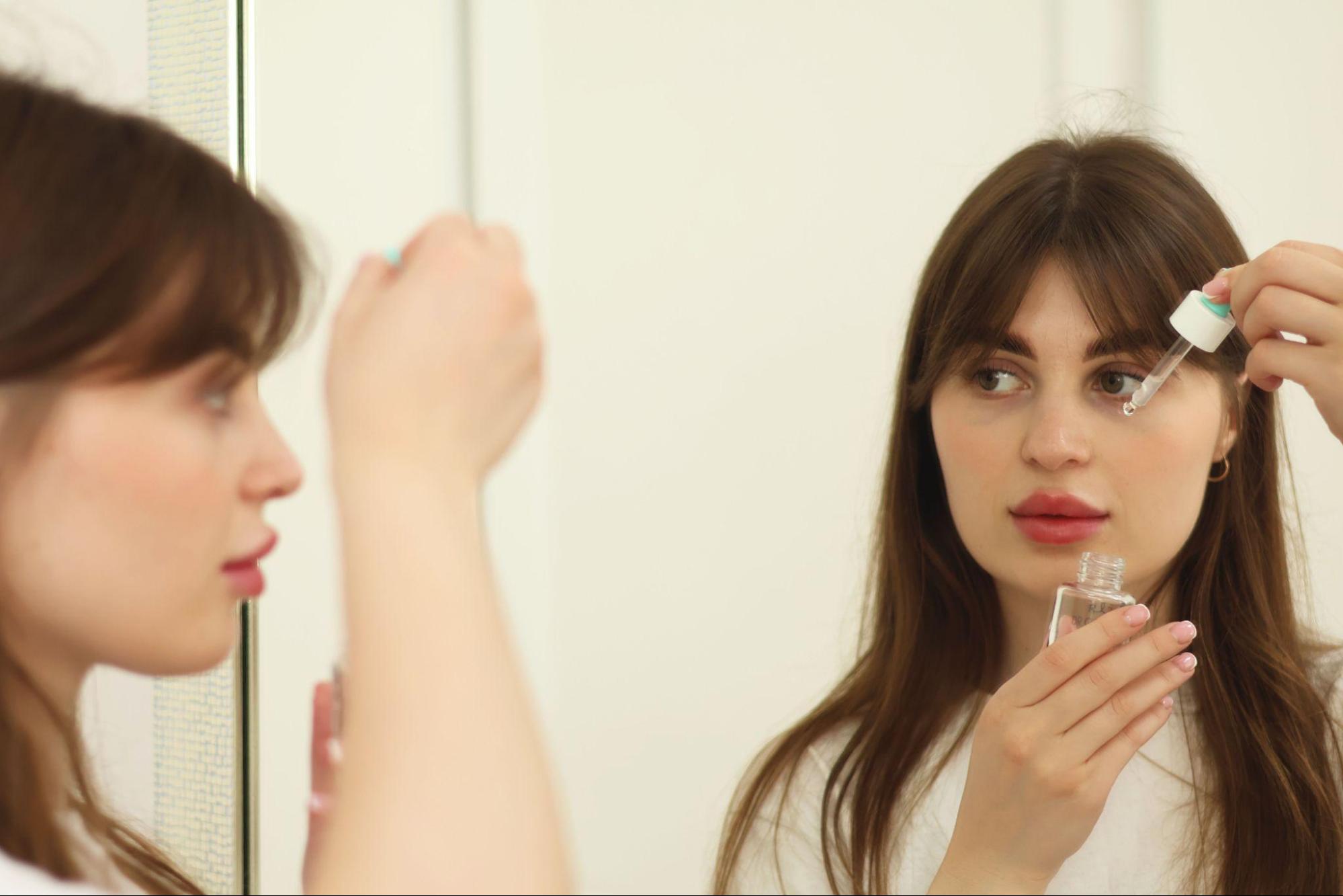 A woman applies makeup in front of a mirror, focusing on her reflection with various cosmetics nearby.