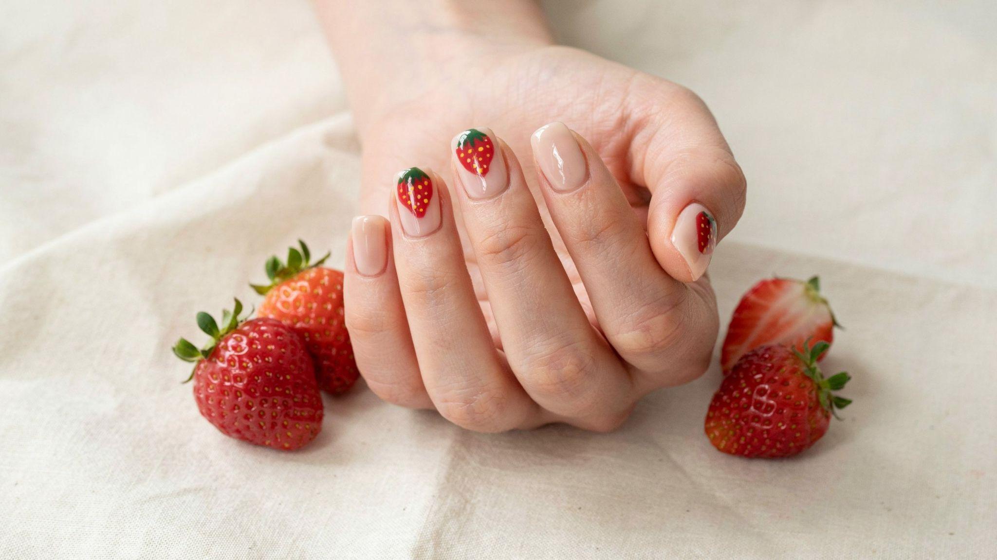 Short square nails featuring adorable strawberry nail art over glossy nude manicure with fresh summer vibes.