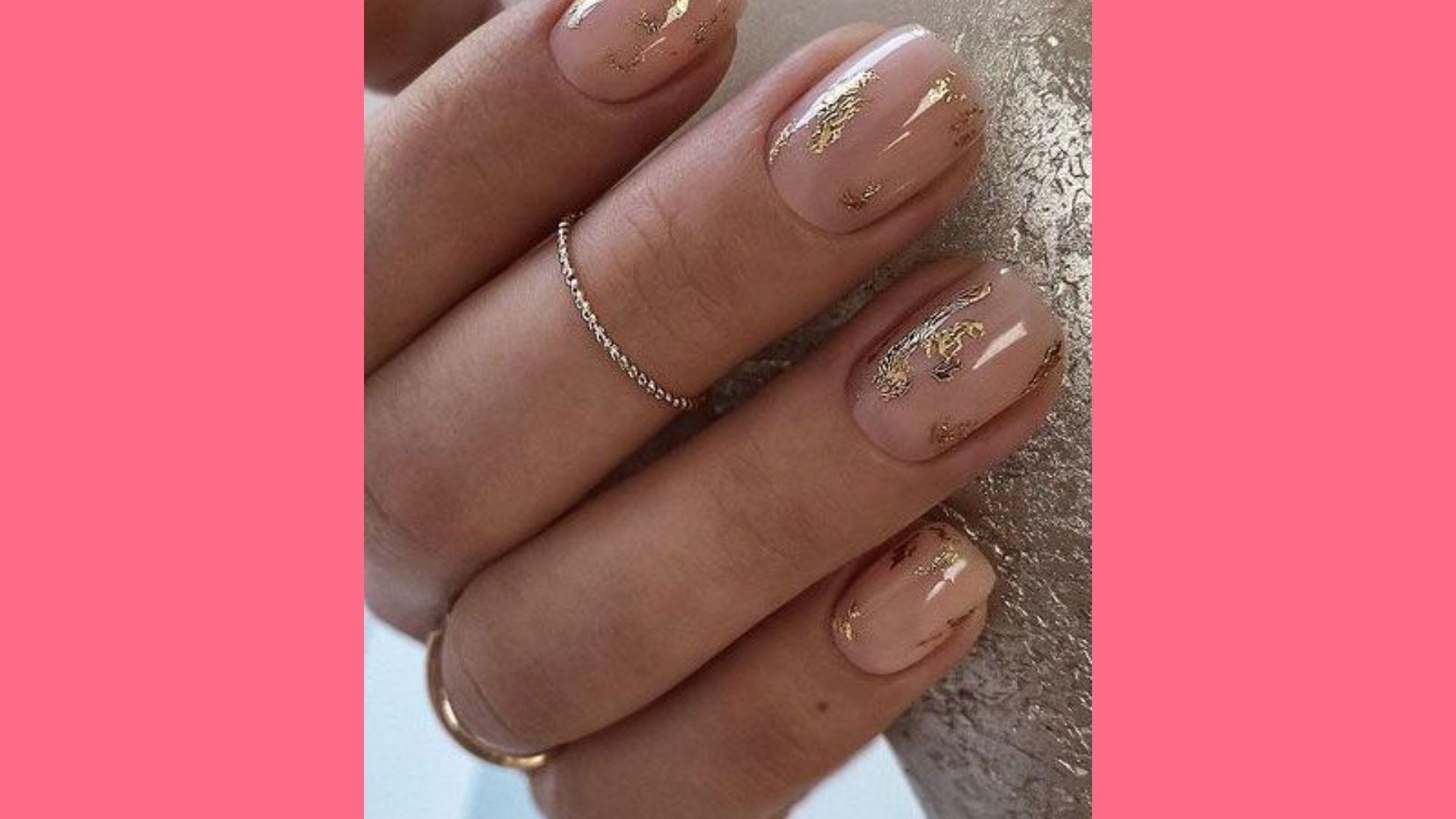 Sheer nude oval nails with gold foil flakes, thin chain ring, gold band, textured gold background