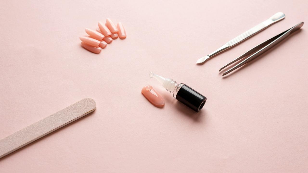 A bottle of nail glue being applied to a press-on nail with manicure tools arranged around it.
