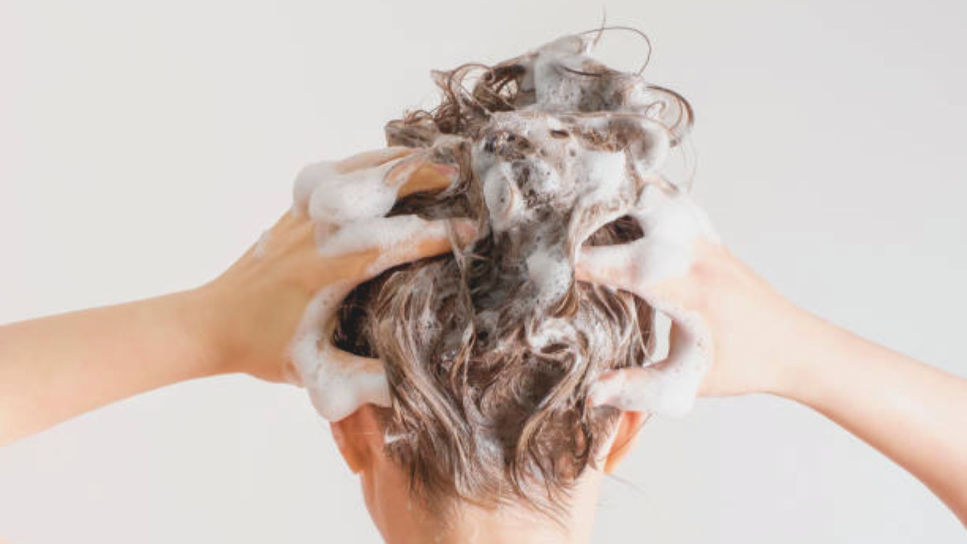 A person washing their blonde hair with soapy lather, viewed from behind.