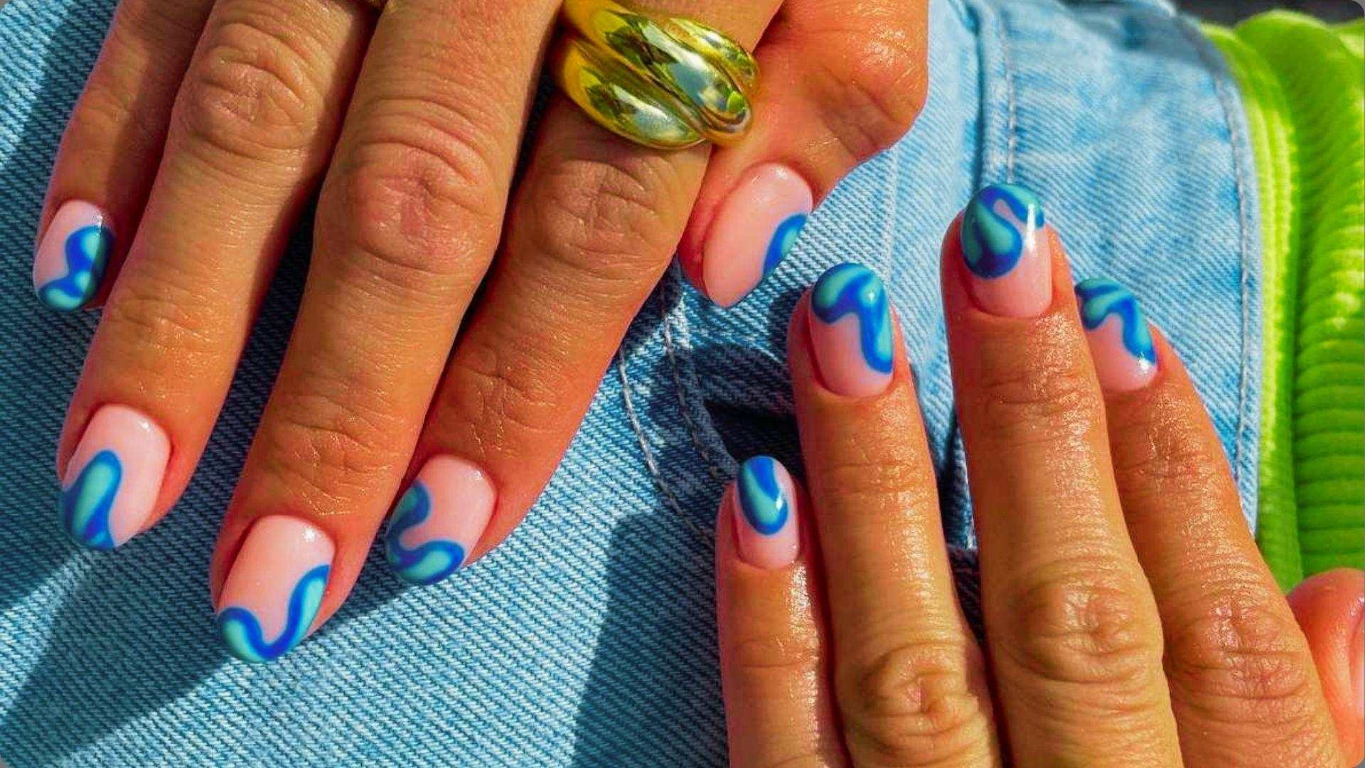 Oval pink nails with blue and mint abstract swirls, chunky gold ring, hands on denim