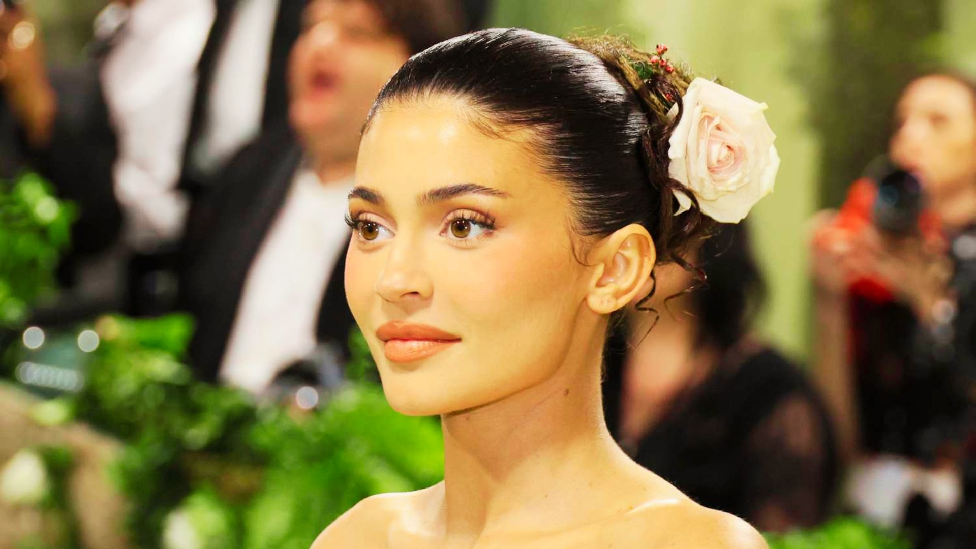 Woman at a formal event with a sleek updo adorned with a pink rose, wearing soft natural makeup and peachy lips.