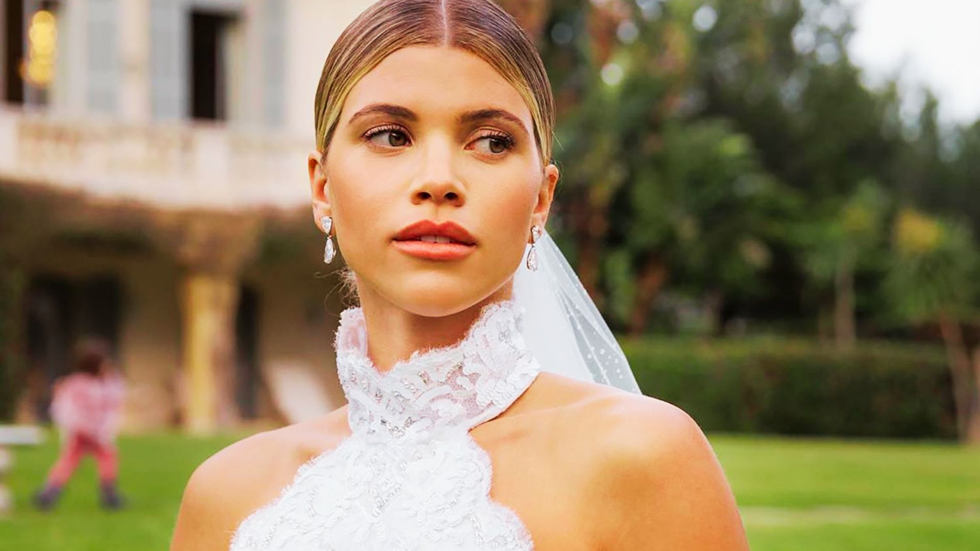 The bride in a high-neck lace gown stands outdoors with a sheer veil and diamond earrings.