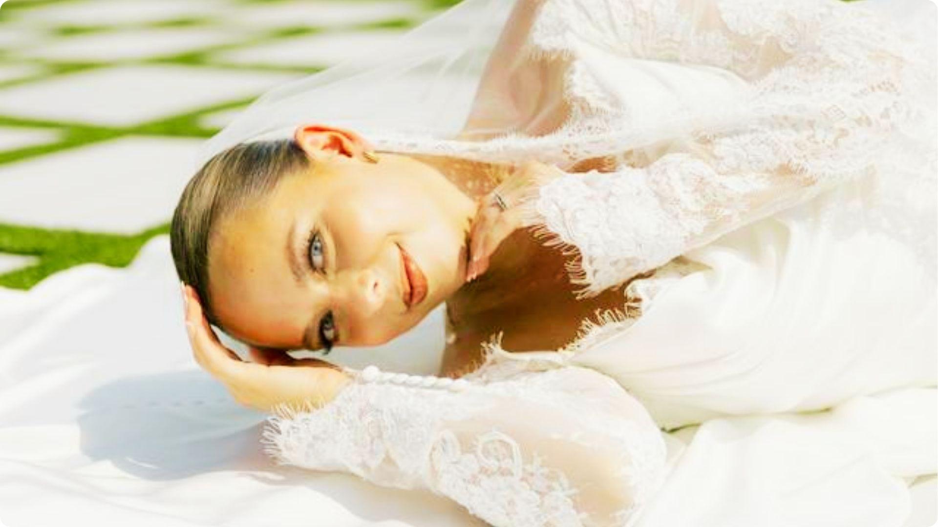Bride in a lace gown and veil lying outdoors, wearing natural wedding makeup with subtle smoky eyes, soft contour, and glossy nude lips.