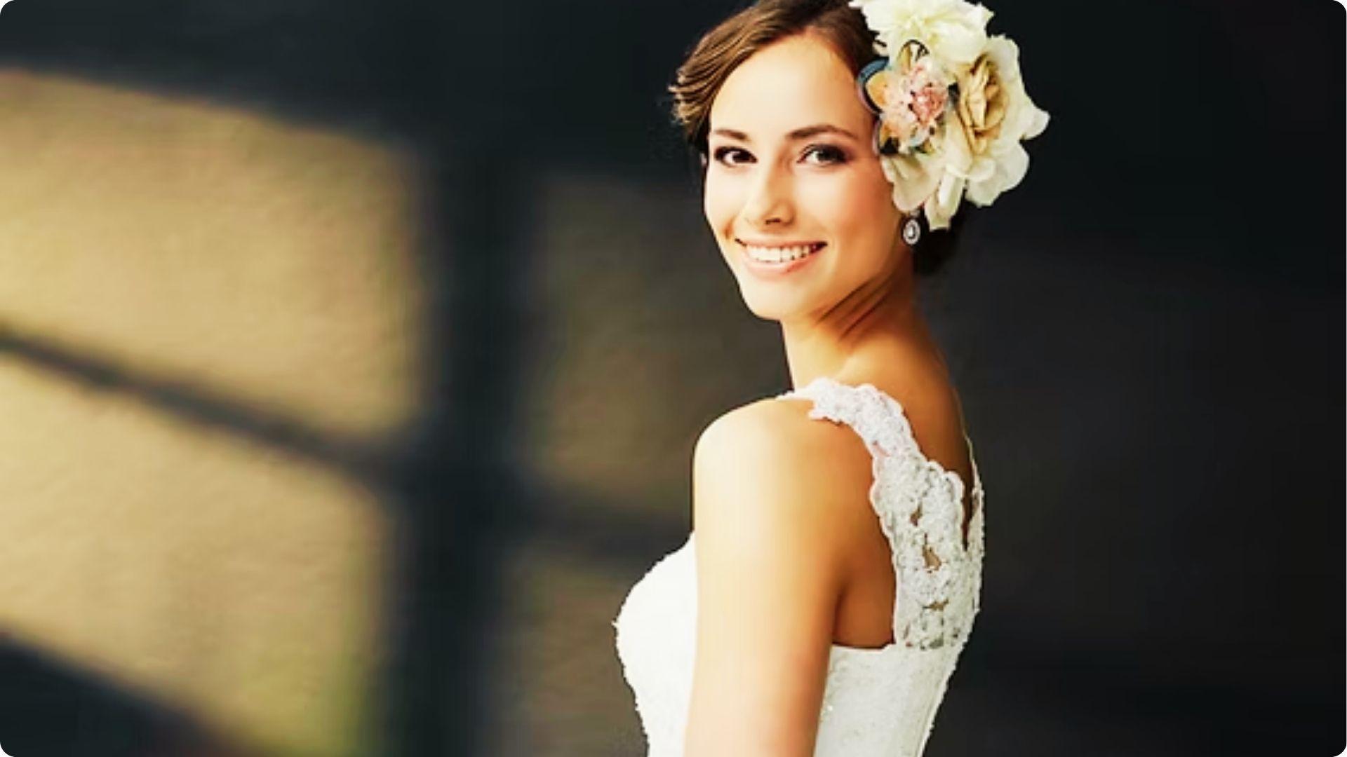 Smiling bride in a lace dress with large floral hairpiece, wearing natural wedding makeup with soft eyeliner, rosy cheeks, and pink lips.