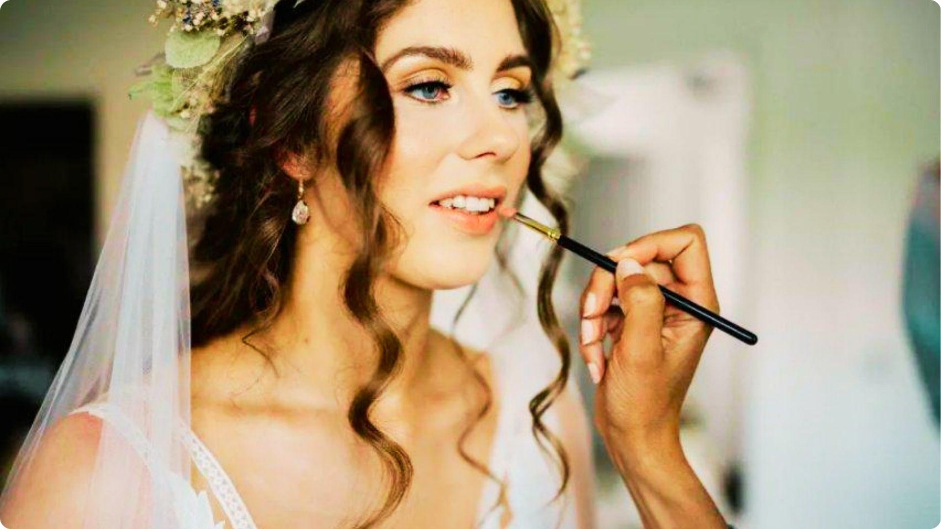 Bride with a floral crown and veil gets lipstick applied during prep, showing soft natural makeup with warm eyeshadow and wispy lashes.