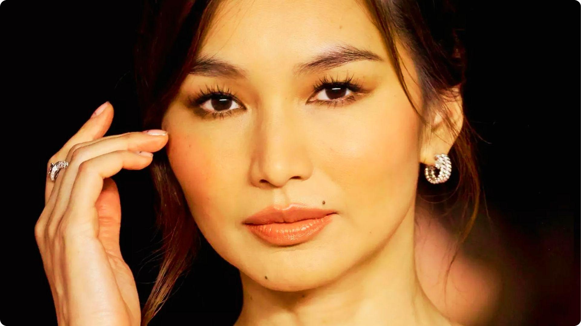 Woman with soft glam makeup featuring glossy coral lips, warm eyeshadow, and dramatic lashes against a black backdrop.