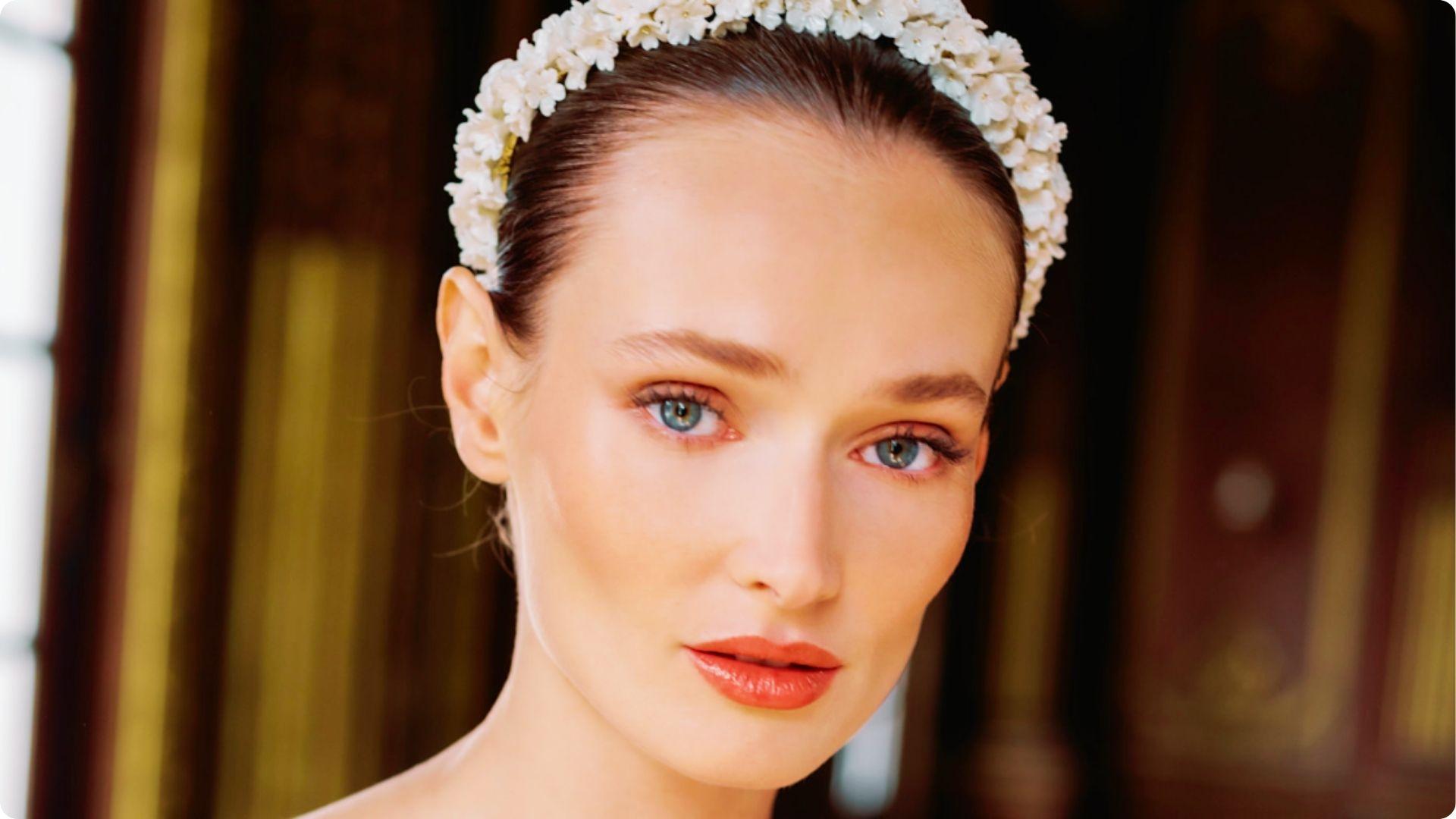Bride with blue eyes wearing a floral crown, featuring soft natural makeup with champagne shimmer, gentle winged liner, and coral gloss.
