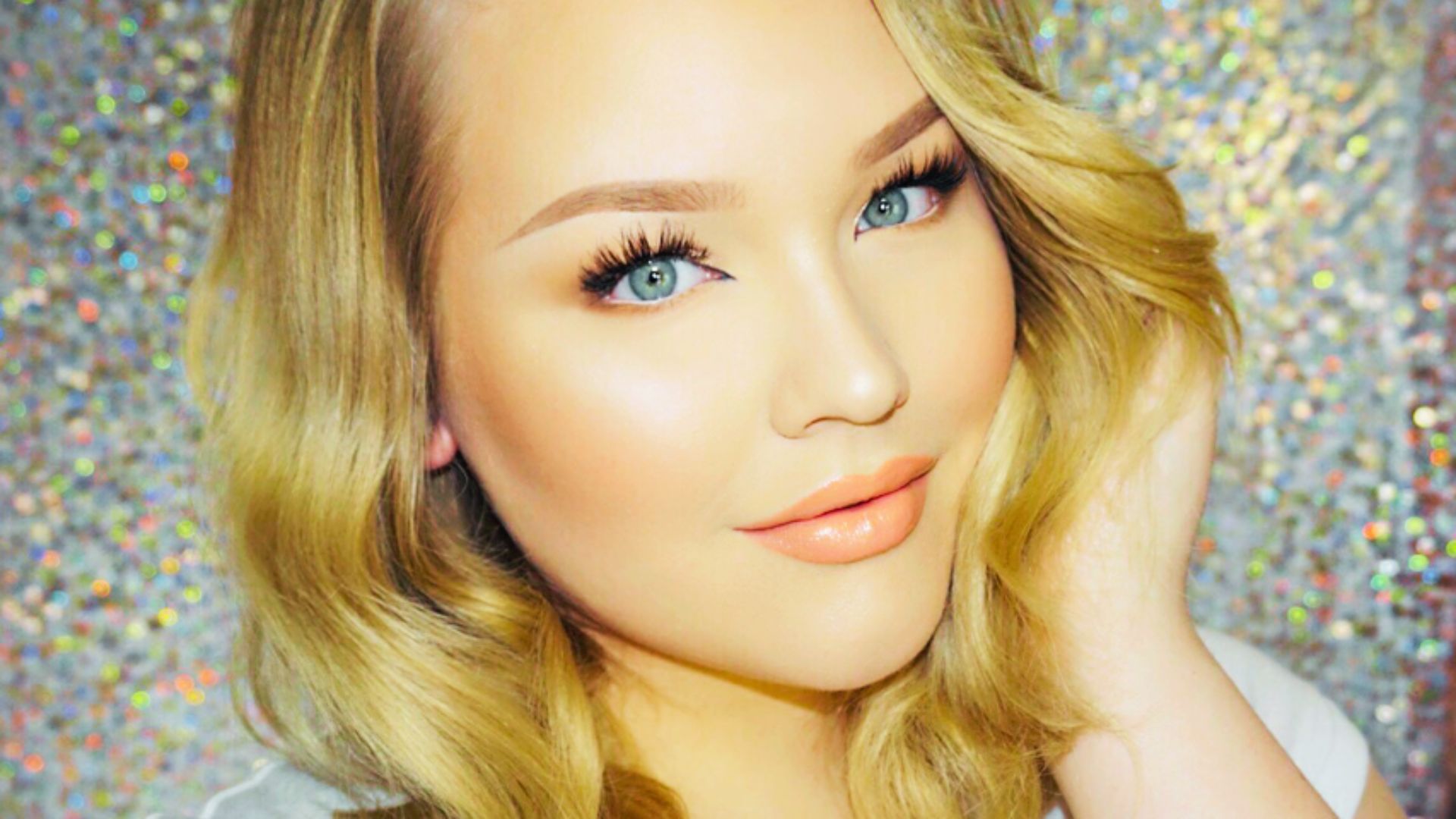Woman with wavy blonde hair and soft glam makeup, including glossy nude lips, fluttery lashes, and radiant skin.