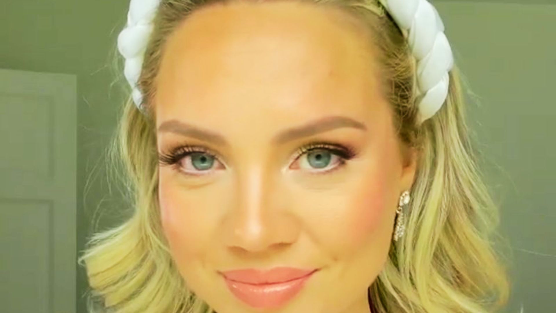 Blonde bride with a braided white headband and diamond earrings smiles softly.